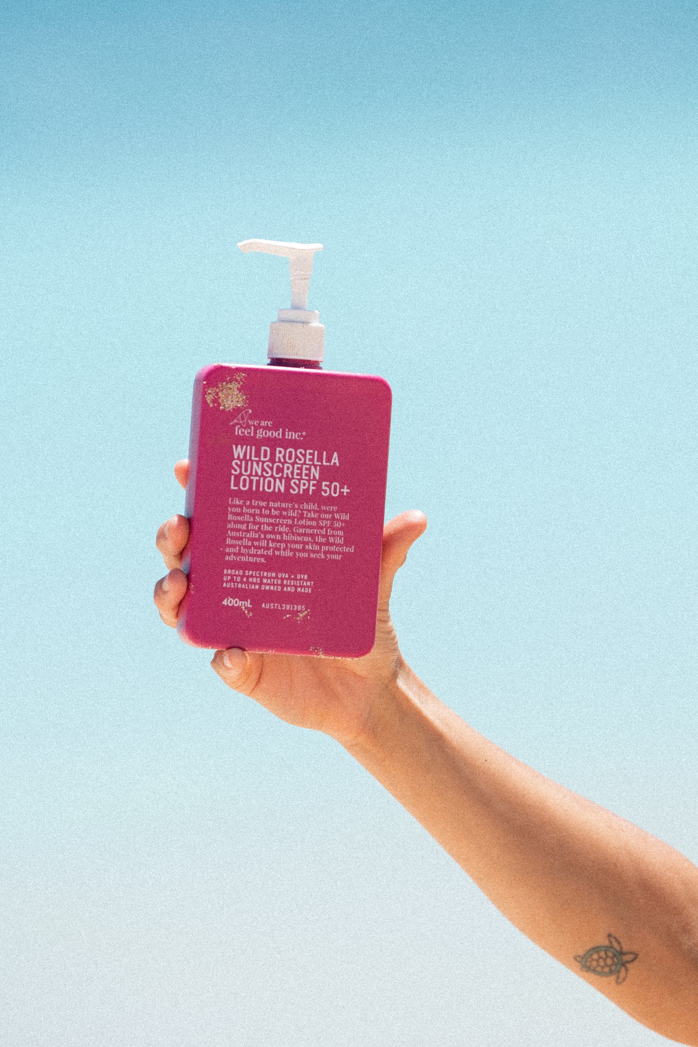 A plastic bottle of We Are Feel Good Inc. Wild Rosella Sunscreen Lotion SPF 50+ held up against a clear blue sky