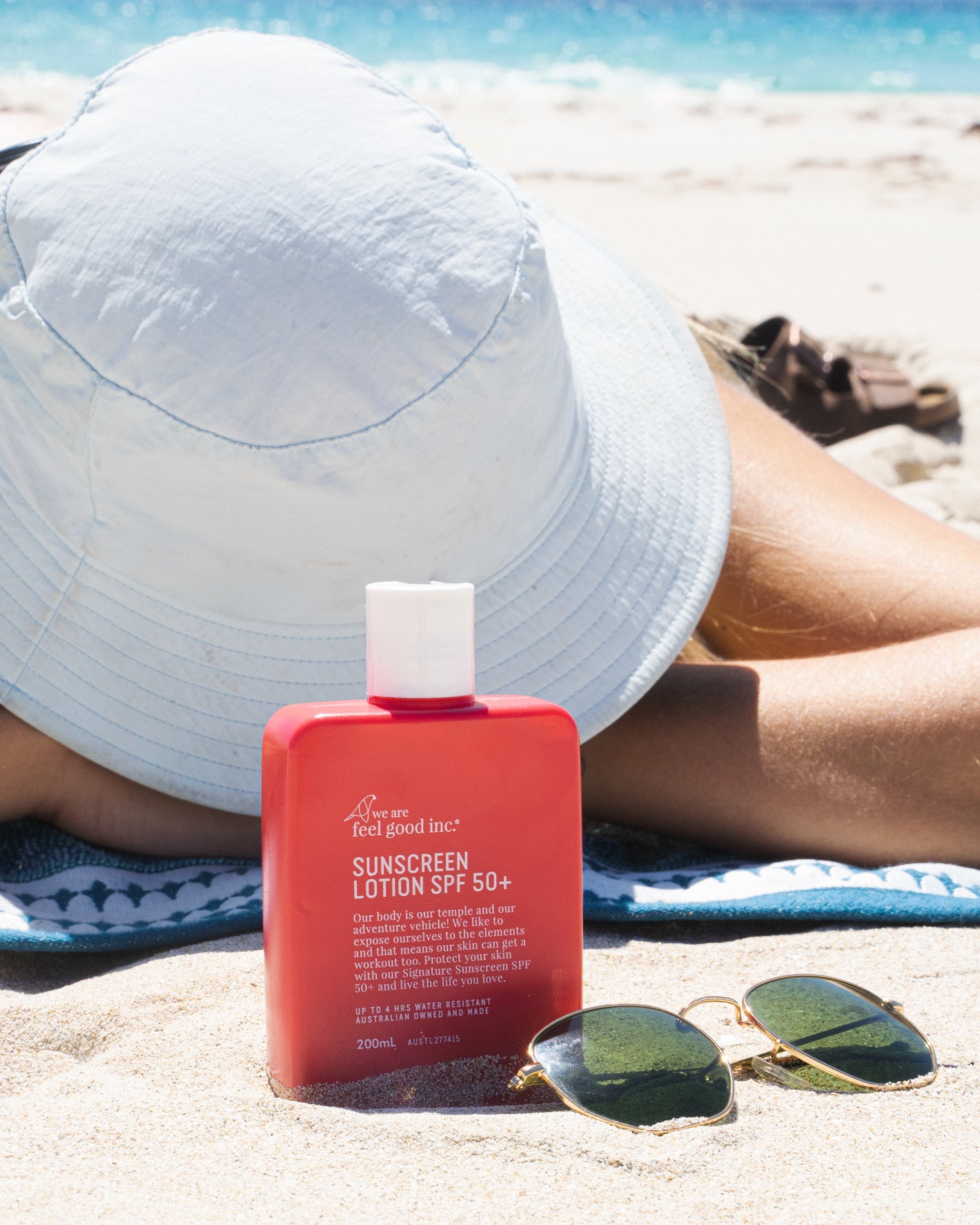 A person lying on the sand wearing a wide-brimmed white hat and sunglasses beside them. In the foreground is a red bottle of 'We Are Feel Good Inc. Signature Sunscreen Lotion SPF 50+' on the sand