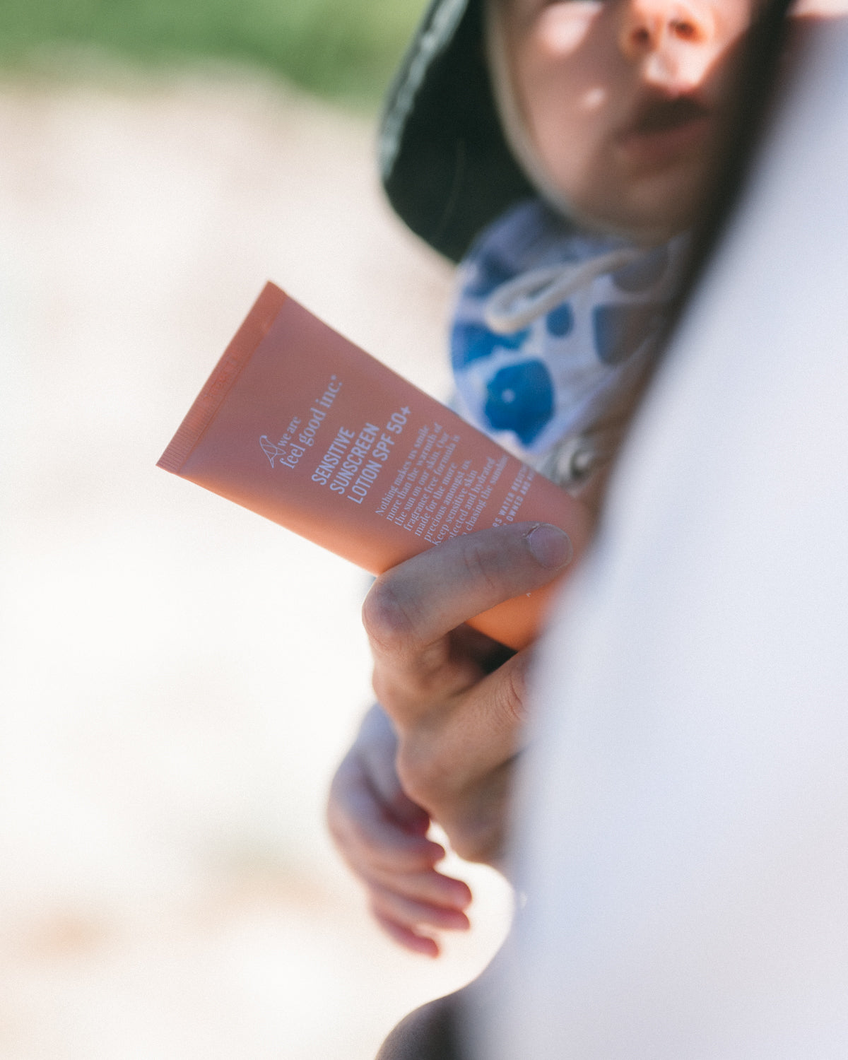 A person's hand is holding an orange tube of We Are Feel Good Inc. Sensitive Sunscreen SPF 50+