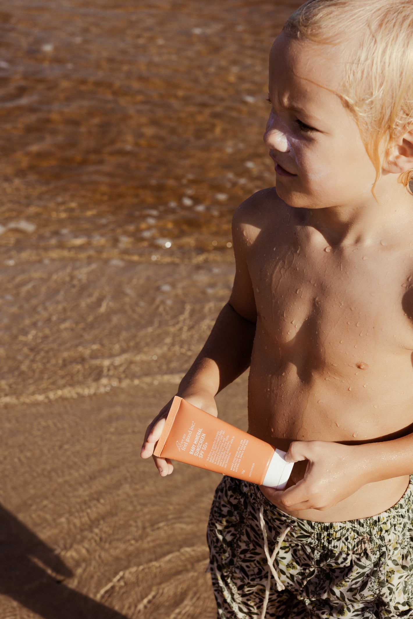 A young child on a sandy beach holding an a tube of We Are Feel Good Inc. Baby Mineral Sunscreen spf50+, with the ocean water lapping near their feet.
