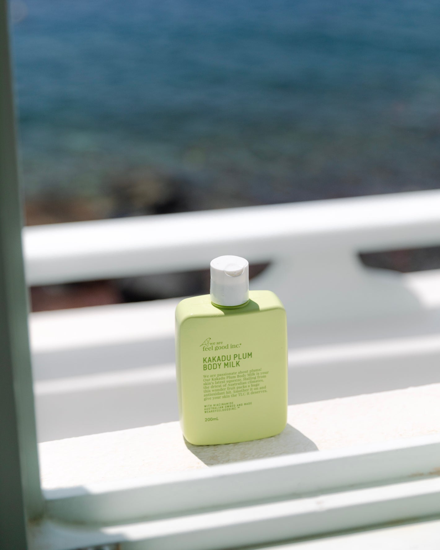 A green bottle of 'We Are Feel Good Inc. Kakadu Plum Body Milk' is placed on a white ledge with a blurred seascape in the background.