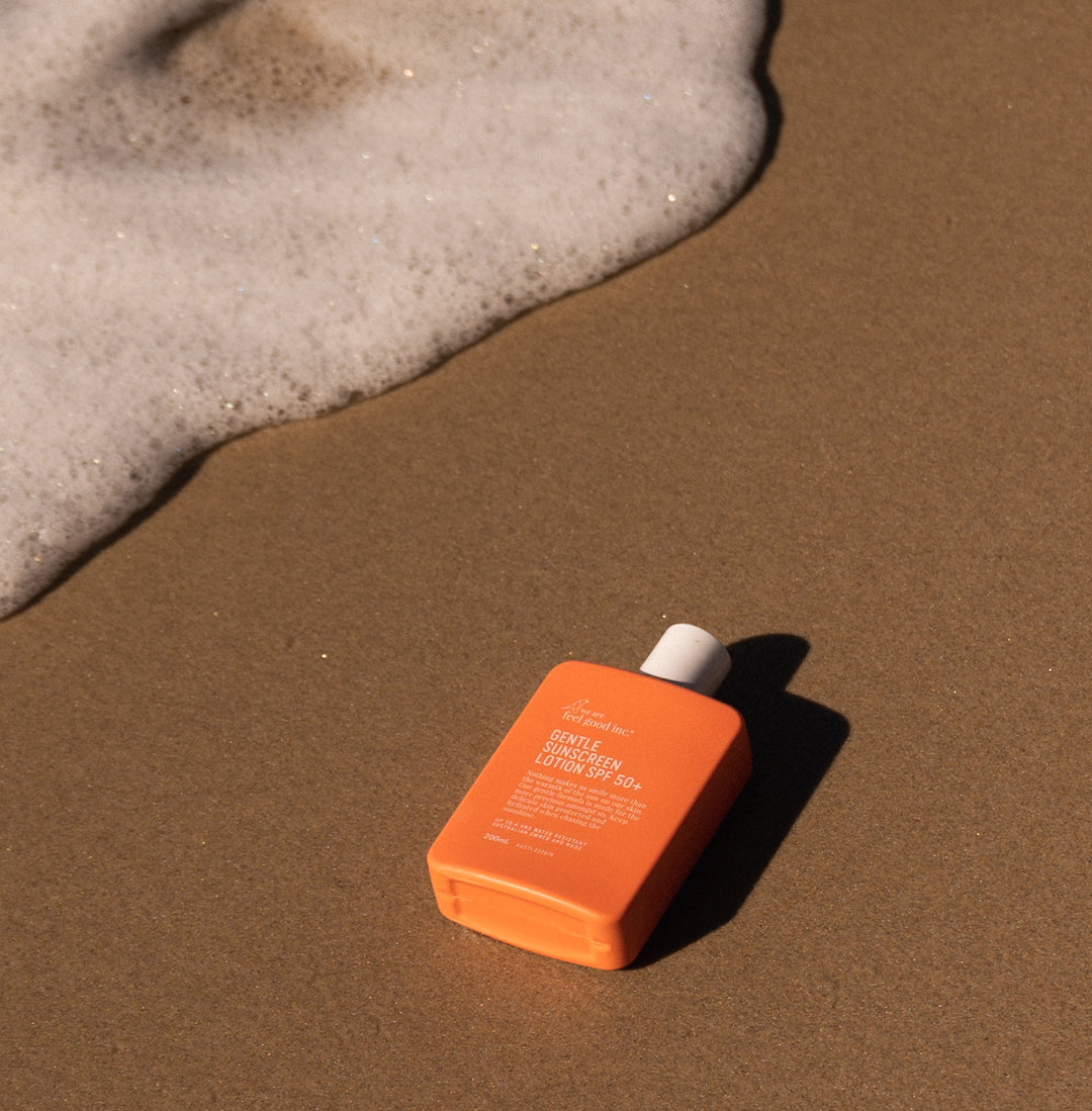 An orange 200ml plastic bottle of We Are Feel Good Inc. Sensitive Sunscreen Lotion SPF 50+ lying on the sand near foamy sea water