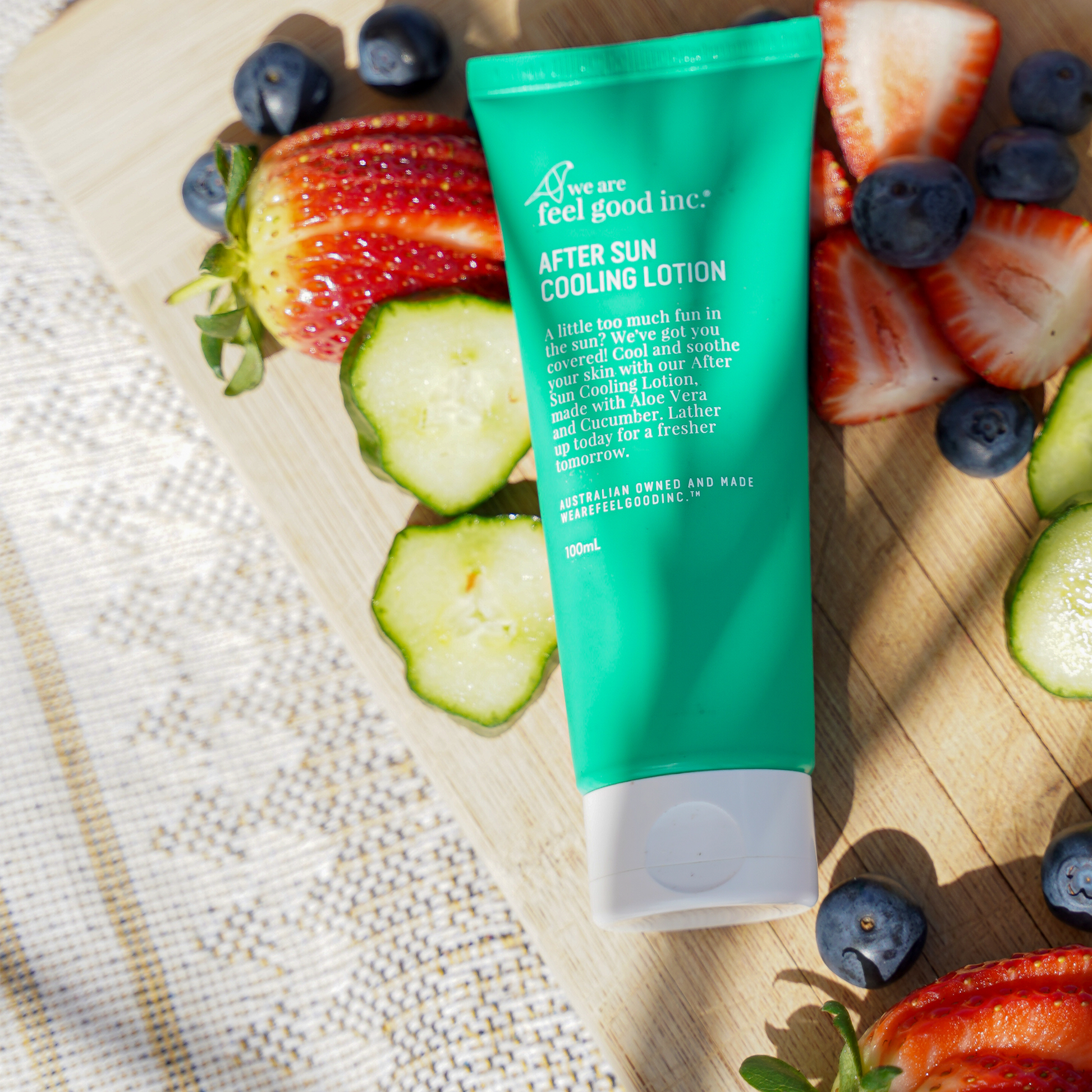 A green tube of 'We Are Feel Good Inc. After Sun Cooling Lotion' is displayed alongside fresh strawberries, blueberries, and cucumber slices on a sunny wooden surface